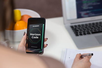 Person holding a phone with 'Horizon Code' course on a desk with a laptop and fruit bowl.